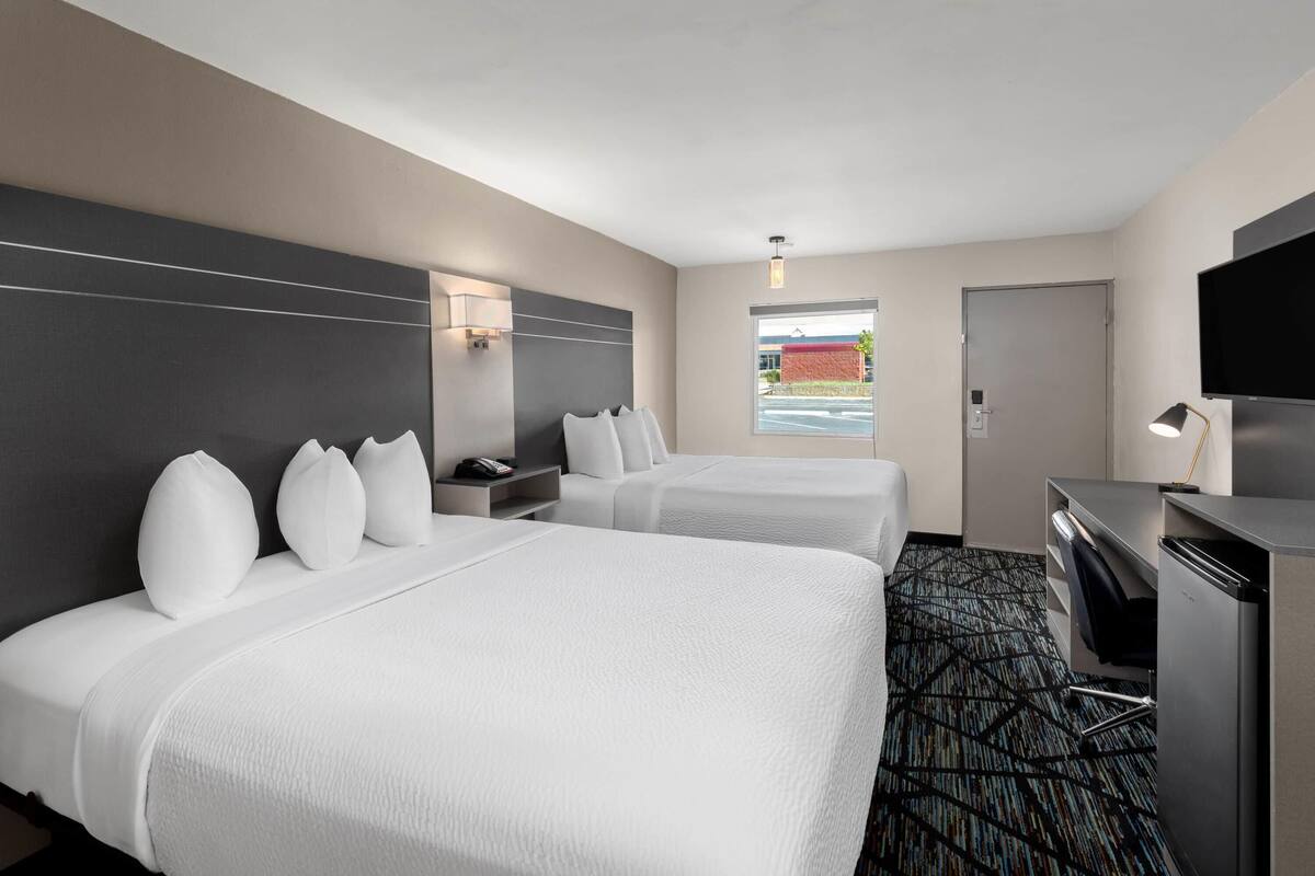 Ett hotellrum med två sängar, ett skrivbord, en tv och ett fönster med utsikt över en byggnad.