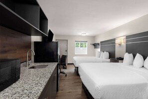 A hotel room with a granite countertop, a sink, a flat-screen TV, two beds, a desk, and a window with a view of trees.