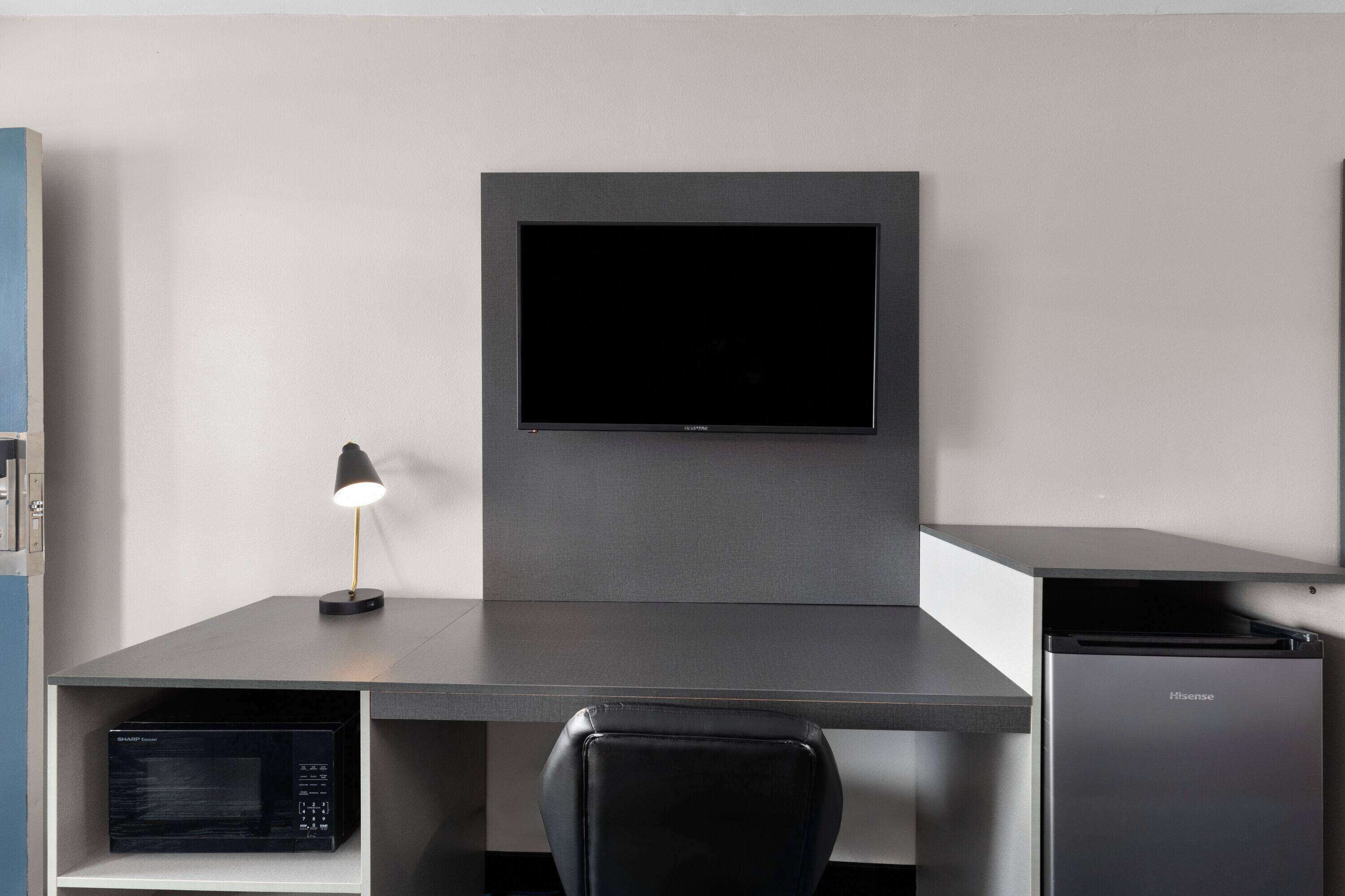 A modern office space with a flat-screen TV mounted on a grey wall, a desk with a lamp, and a black chair.
