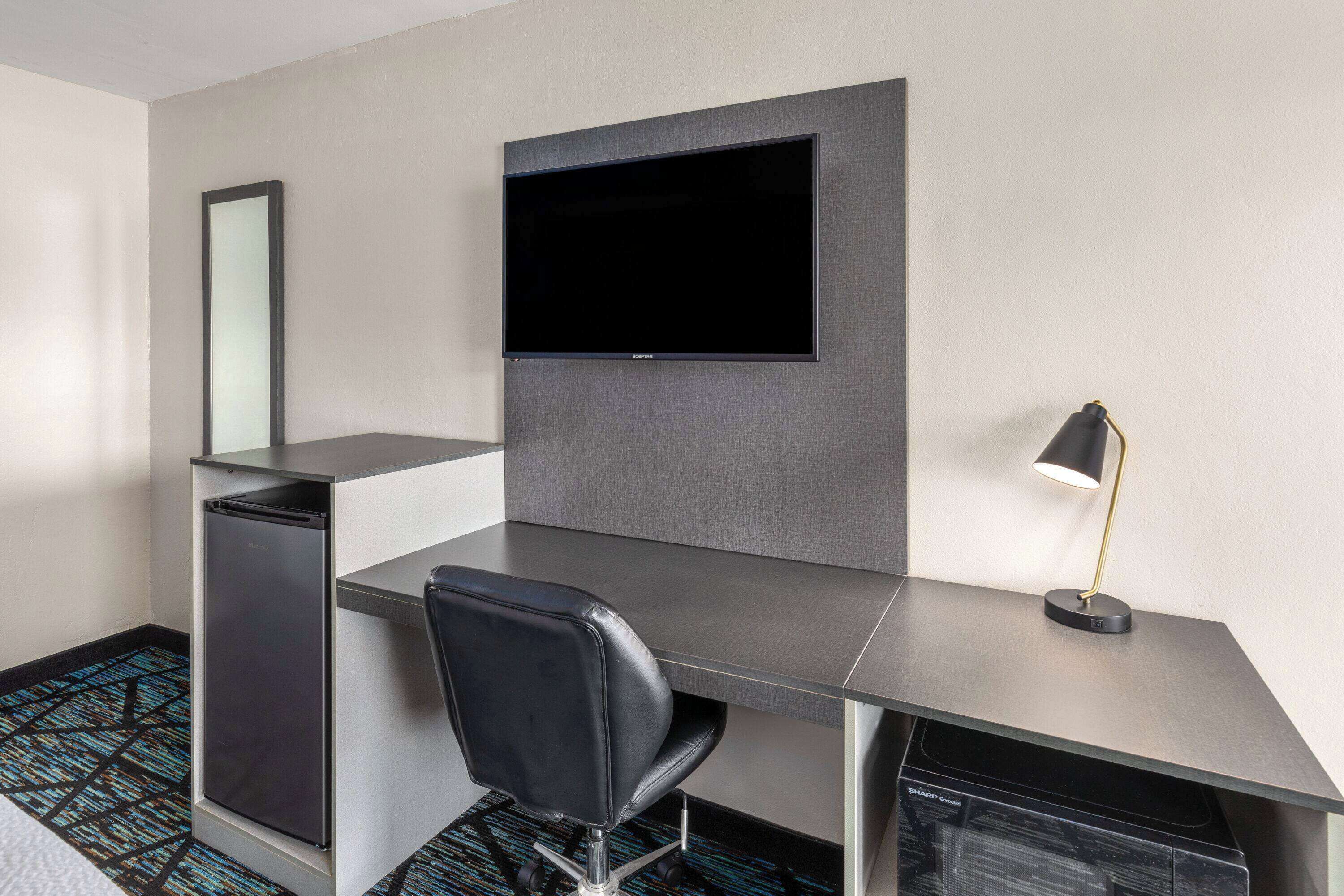 Habitació d'hotel moderna amb televisió de pantalla plana penjada a la paret, escriptori amb cadira, nevera petita i làmpada d'escriptori.