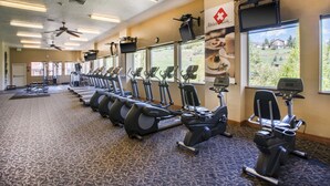 Un gimnasio con varias cintas de correr y bicicletas estáticas, ventiladores de techo y amplias ventanas con vistas al exterior.