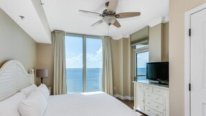Un cuarto de hotel con una cama grande, un ventilador de techo, un televisor sobre un tocador y vista al océano a través de ventanales.