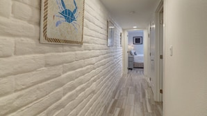 Un pasillo con paredes de ladrillo blanco, una obra enmarcada de un cangrejo azul y una cama con cabecera blanca al fondo.