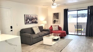 Un salon moderne avec un canapé gris, un fauteuil rouge, une table basse en verre et une grande fenêtre avec des rideaux.
