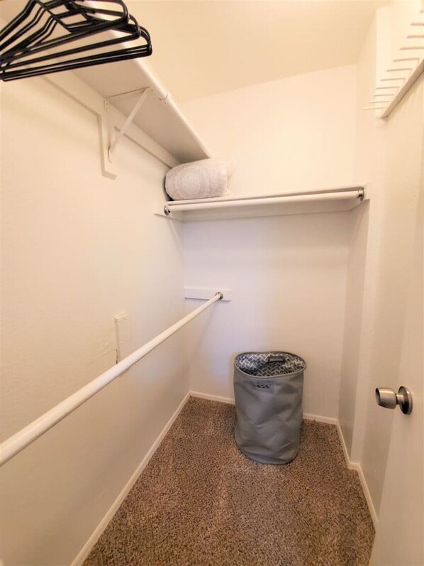 Un coin de pièce avec un mur blanc, une moquette beige, une porte blanche, une poubelle et un porte-manteau noir.