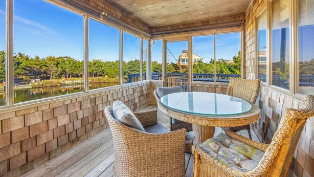 Eine überdachte Terrasse mit Korbmöbeln, großen Fenstern und Blick auf einen See sowie Gebäude.