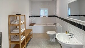 A modern bathroom with a bathtub, toilet, and sink. There is a wooden shelf with towels and a window with blinds. - Yew Tree Barn - HW7737 (Near Usk)