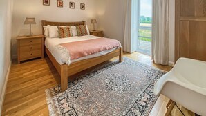 A bedroom with a wooden bed, a nightstand, a bedside table, a patterned rug, and a view of the outdoors through a window. - Yew Tree Barn - HW7737 (Near Usk)