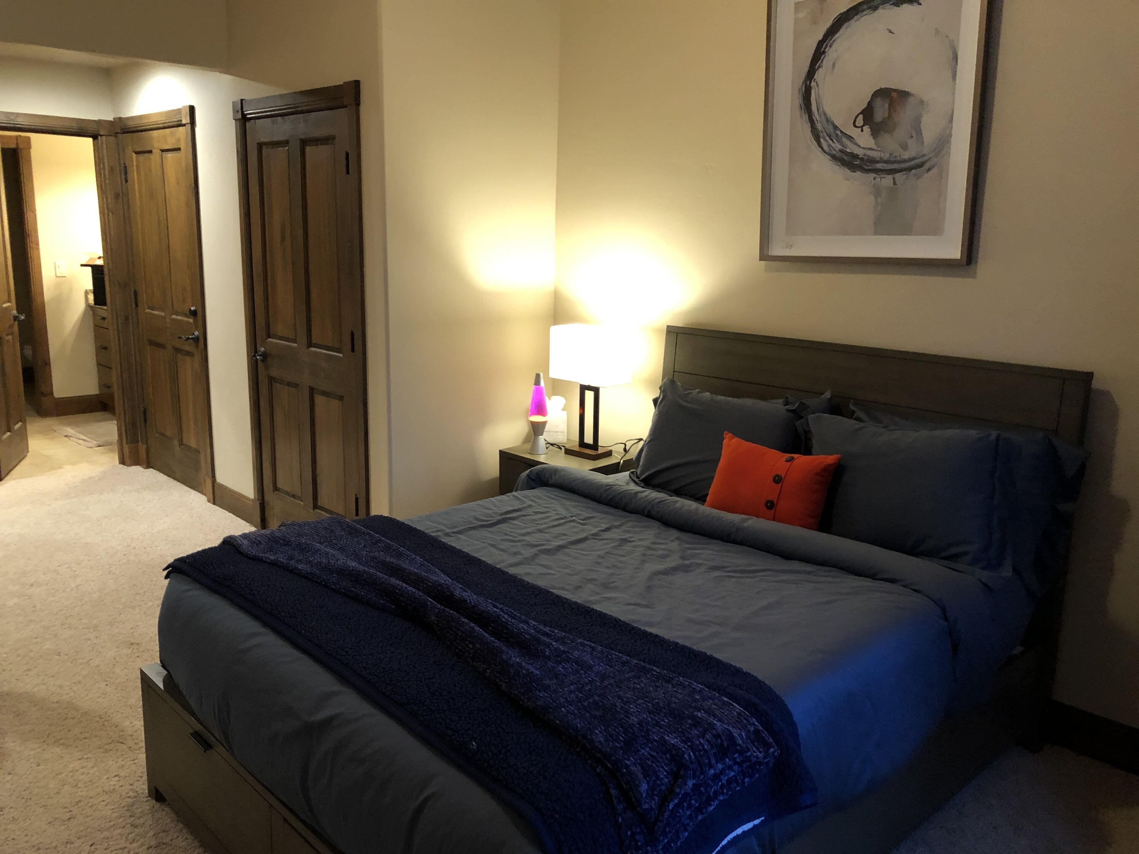 A bedroom with a bed, nightstand, lamp, and artwork on the wall.