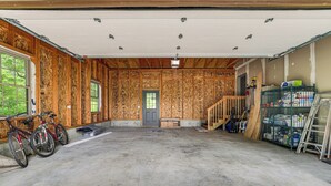 Een garage met houten wanden, een betonnen vloer en een deur naar buiten. Tegen de muur staan twee fietsen geparkeerd en er is diverse gereedschap en apparatuur in de hoek opgeborgen.