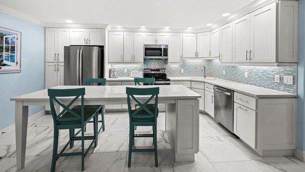 A modern kitchen with white cabinetry, a stainless steel refrigerator, a microwave, an oven, and a dishwasher. There are two green chairs at a white table. - Cozy Dockside 2 bed, 2 bath at Sanibel Moorings #1422 (Sanibel)