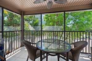 Eine überdachte Terrasse mit einem runden Glastisch und Korbstühlen, umgeben von einem Metallgeländer und Glasscheiben.