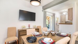 Zona pranzo con tavolo in legno apparecchiato per quattro persone, televisione a parete e vista sull'esterno attraverso una finestra.
