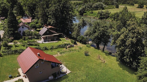 Una casa di campagna con tetto rosso, immersa nel verde e bagnata da un fiume.