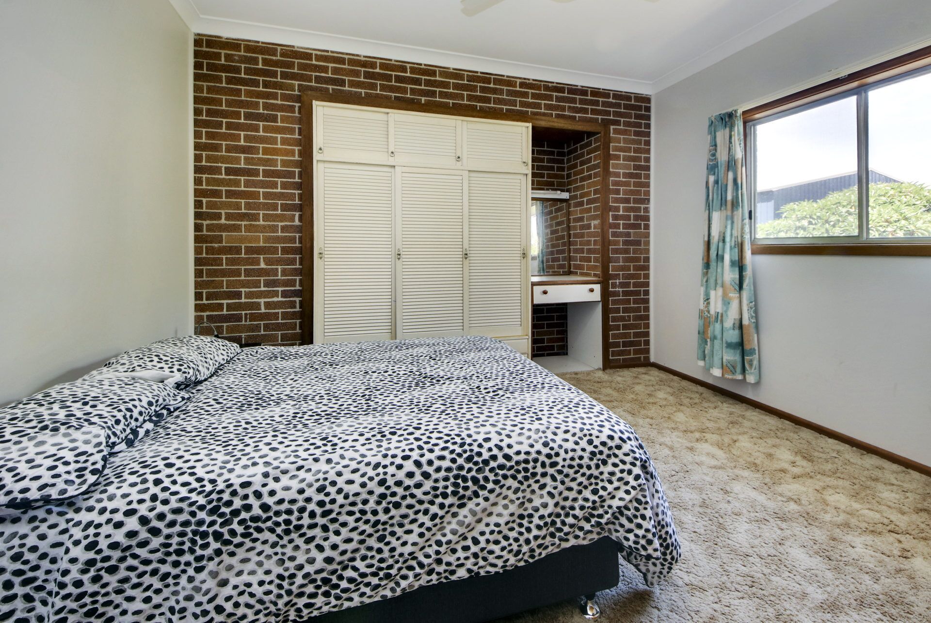 A bedroom with a brick wall, a bed with a leopard print bedspread, a white wardrobe, and a window with curtains.