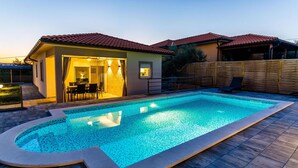 Ein Schwimmbad mit gefliestem Sonnendeck, eine überdachte Terrasse mit Gartenmöbeln und ein Holzzaun.