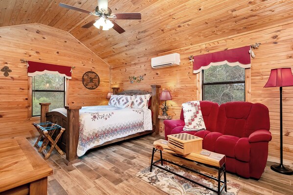 L’intérieur d’un chalet accueillant, avec un plafond en bois, un lit recouvert d’une courtepointe à motifs floraux, un fauteuil rouge, une petite table en bois et un ventilateur de plafond.