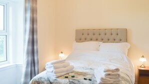 Una cama bien tendida con toallas dobladas, una mesita de noche con una lámpara y una ventana con cortinas.