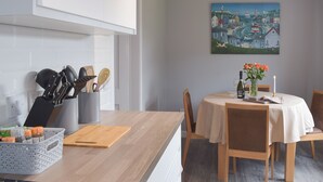 Una cocina moderna con armarios blancos, encimera de madera y un comedor con mesa y sillas redondas. En la pared, un cuadro que representa una escena costera.