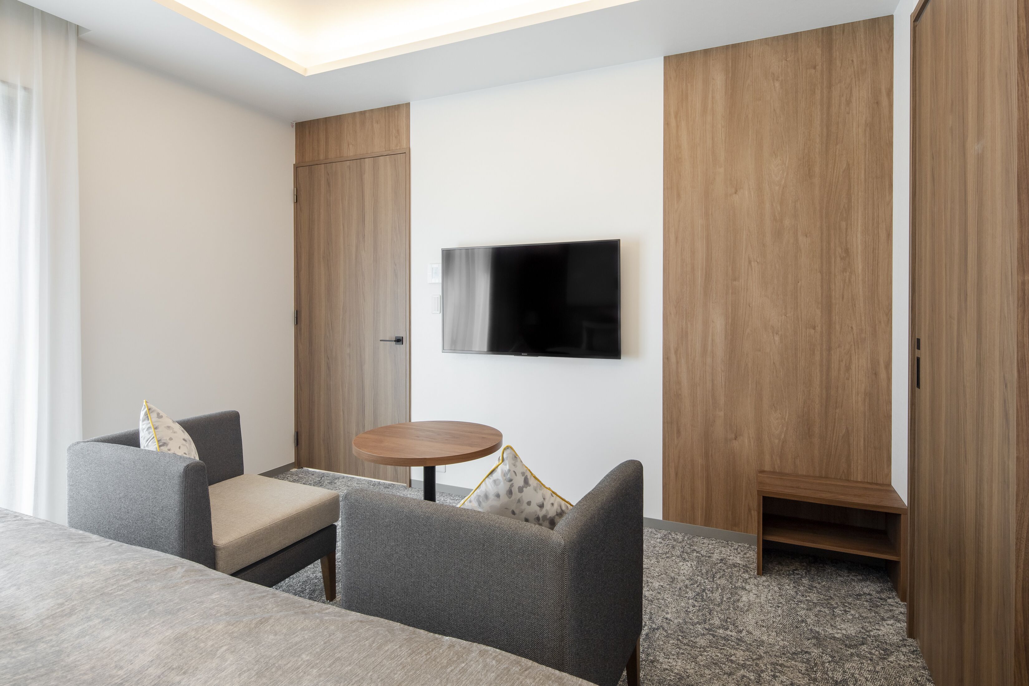 Una habitación de hotel moderna con un televisor de pantalla plana montado en la pared, un sofá gris con un cojín, una pequeña mesa de madera y un armario empotrado.