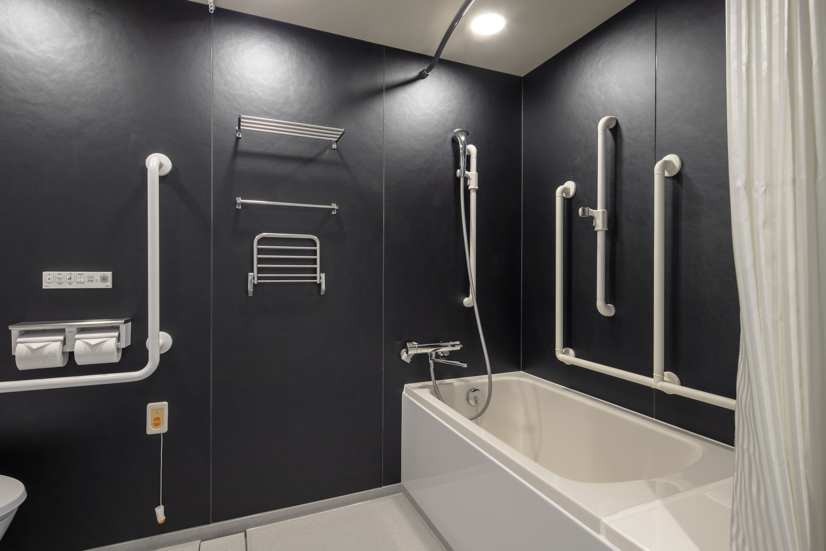 A modern bathroom with a bathtub, handrails, a towel rack, and a toilet paper dispenser.