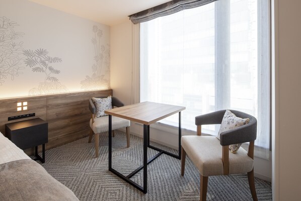 A modern room with a wooden bedside table, a small bedside table with a lamp, a chair with a cushion, and a large window with sheer curtains.