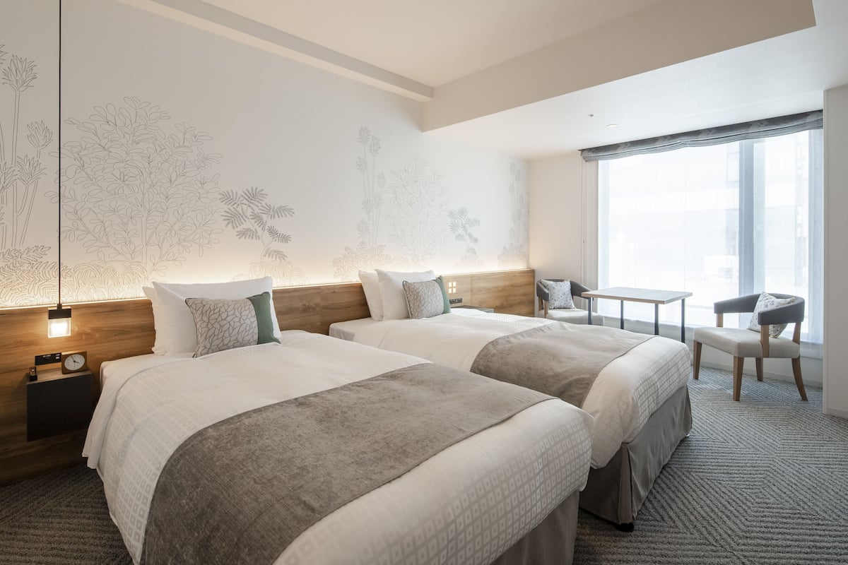 A hotel room with two beds, a small table, a chair, and a large window.