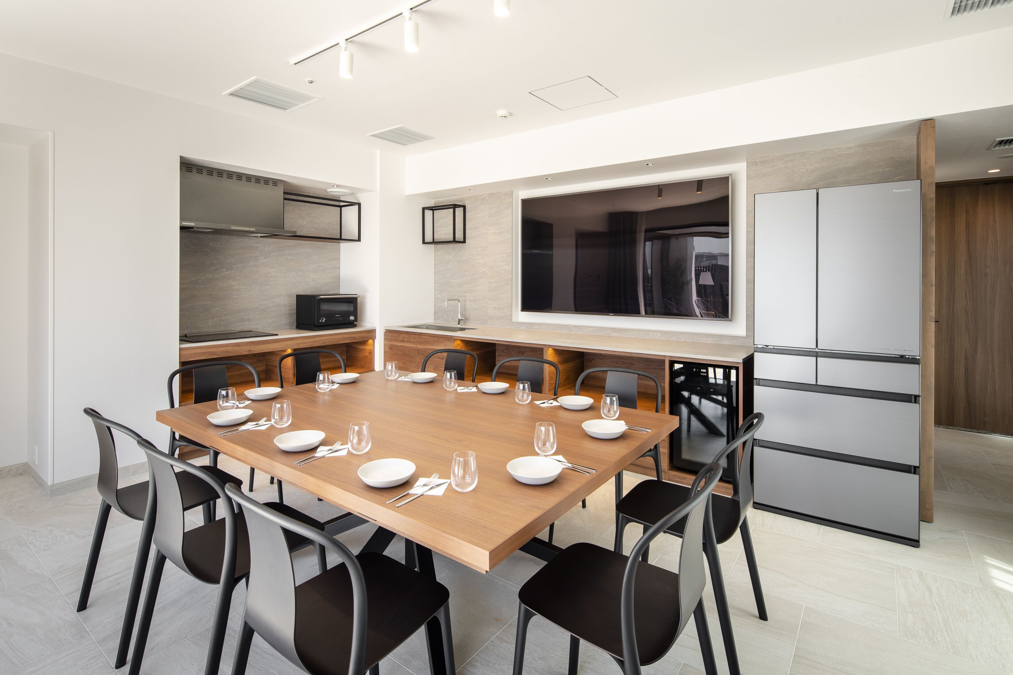 A modern dining area with a wooden table set for six, surrounded by black chairs. A kitchen with a large window and a refrigerator is visible in the background.