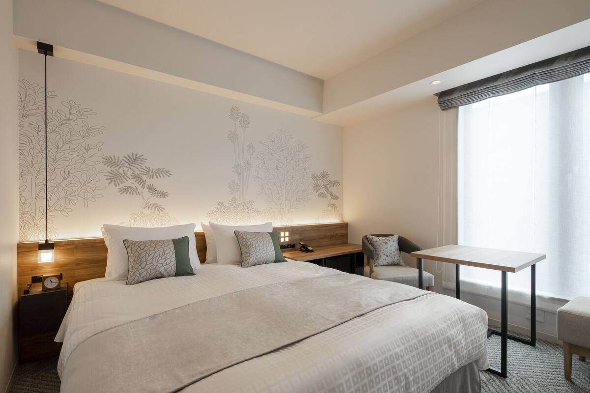 A modern hotel room with a large bed, a desk, and a chair. There is a nightstand with a clock and a lamp. The room has a large window with curtains.