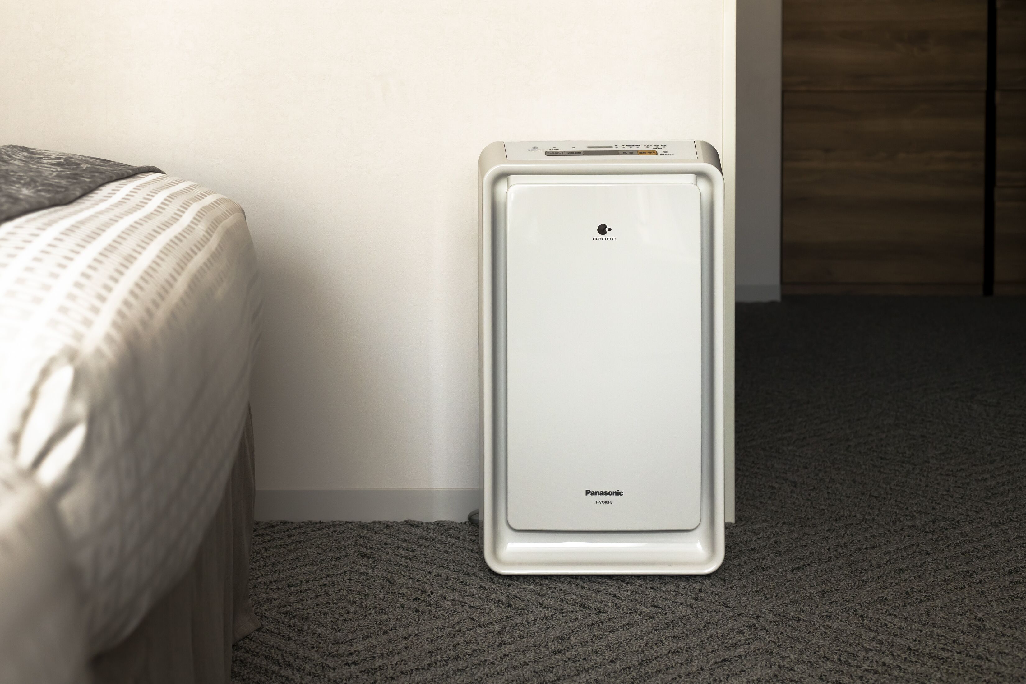 Un purificador de aire Panasonic blanco en una habitación con suelo alfombrado y una cama con edredón estampado.