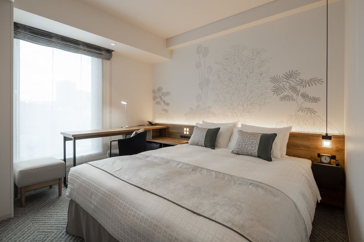 A modern hotel room with a large bed, a desk, and a chair. There is a wall-mounted clock and a bedside table with a lamp.