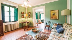 Una sala de estar con sofá, mesa, silla, radiador y ventana con contraventanas verdes.