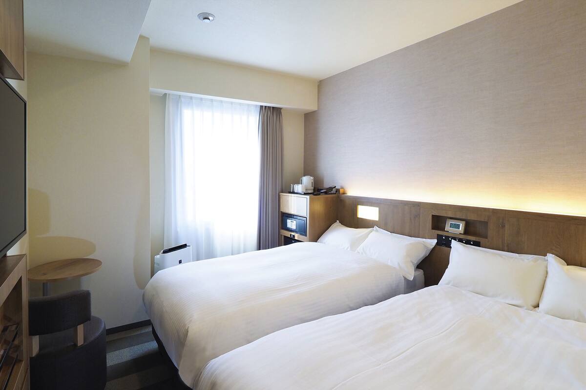 A hotel room with two beds, a desk, a TV, and a window with curtains.