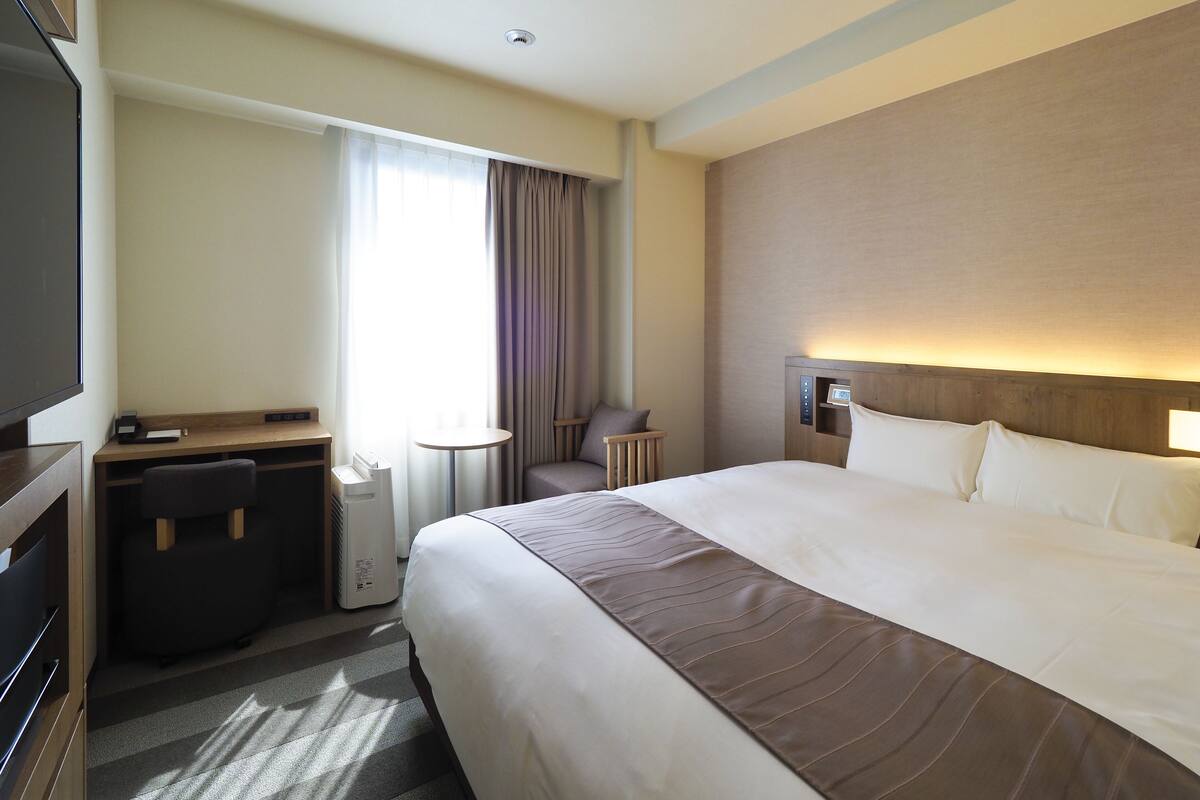 A hotel room with a large bed, a desk, a chair, a TV, and a window with curtains.