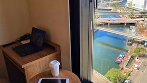 A room with a wooden desk, a laptop, a tablet, and a coffee cup. A large window offers a view of a river with boats and a bridge. - Far East Village Hotel Yokohama (Yokohama)