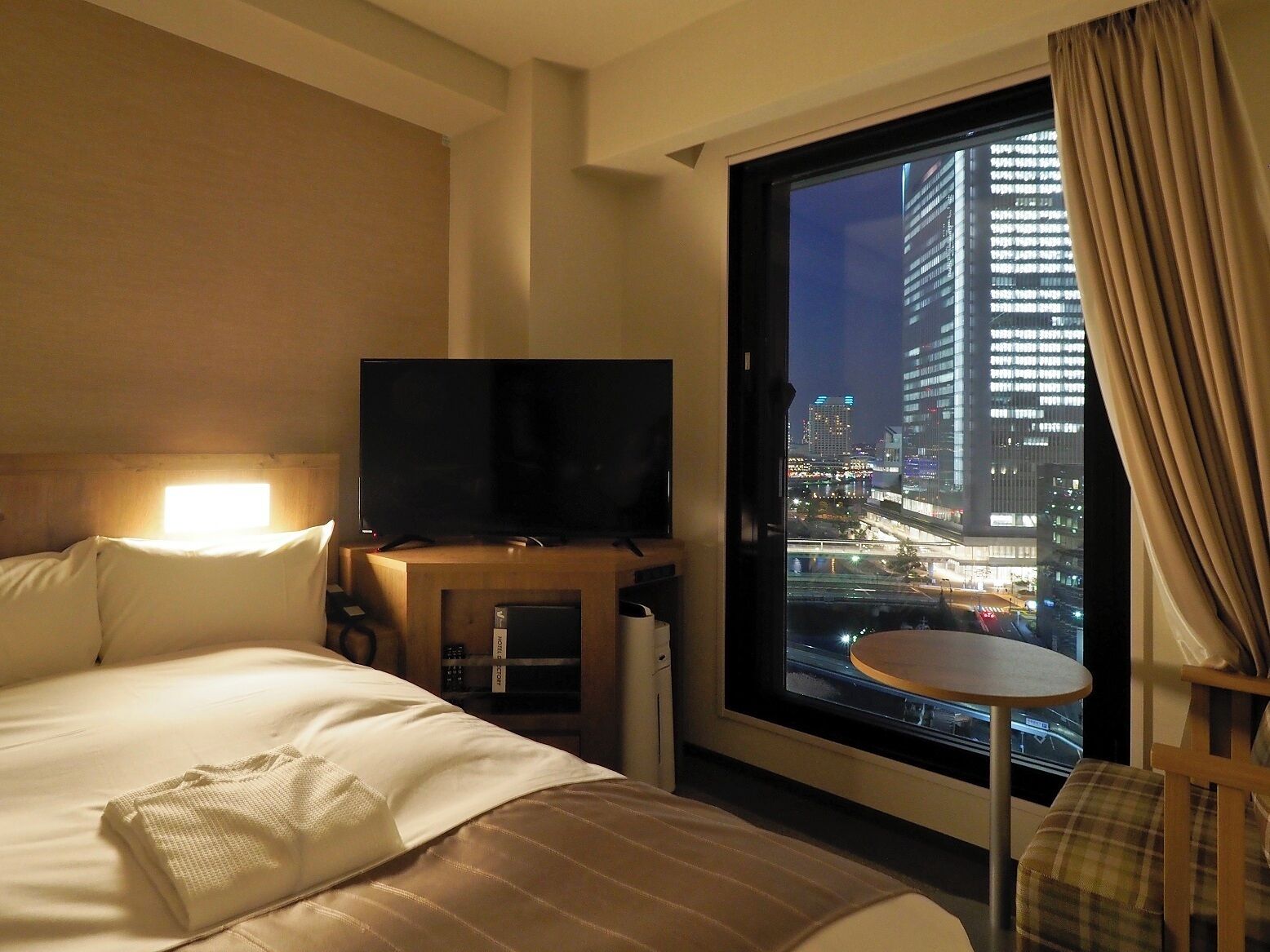 Une chambre d’hôtel avec un lit, un téléviseur posé sur un meuble en bois, une petite table et une vue sur le paysage urbain illuminé la nuit.