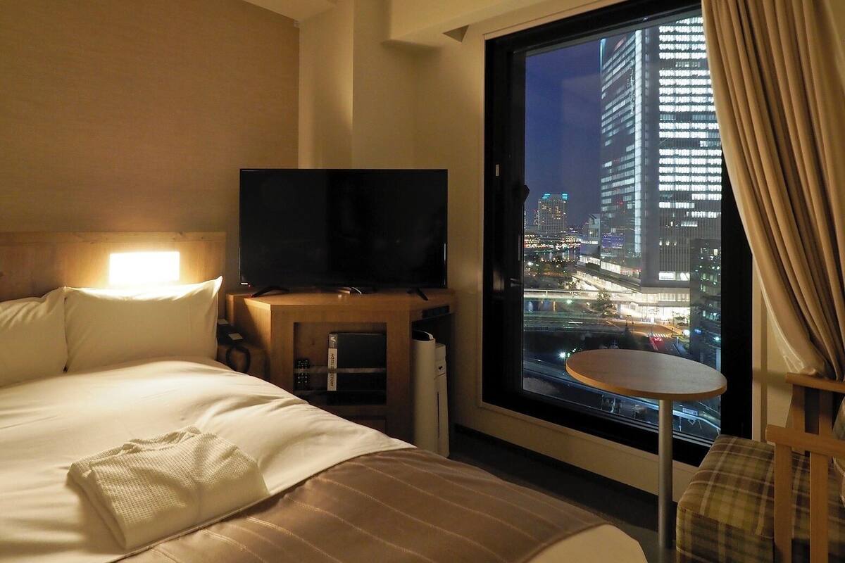A hotel room with a bed, a television on a wooden stand, a small table, and a view of a cityscape at night.