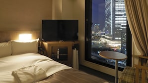 A hotel room with a bed, a TV, a small table, and a view of a cityscape at night. - Far East Village Hotel Yokohama (Yokohama)