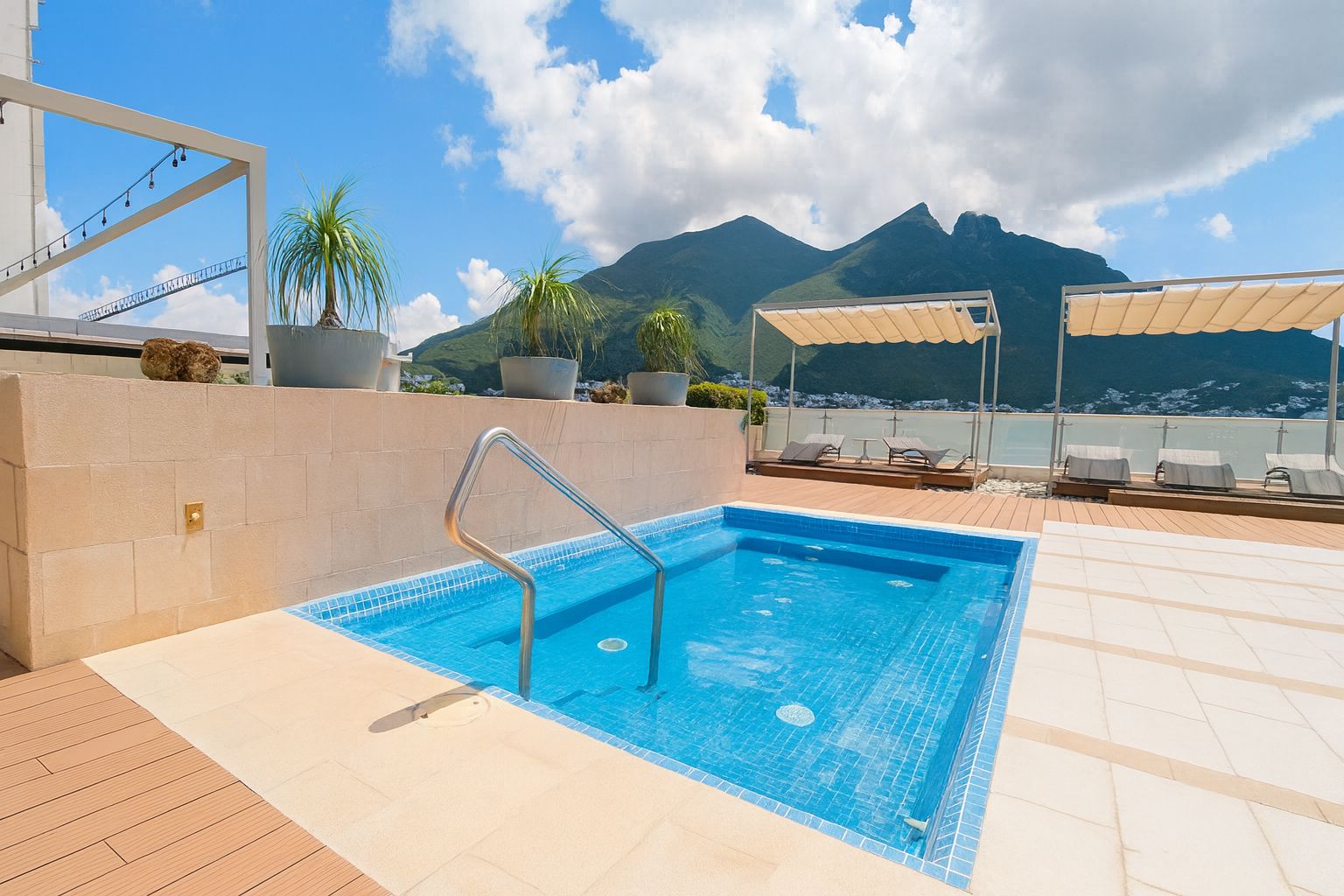 A rooftop pool with a clear view of mountains in the background, surrounded by lounge chairs and potted plants.