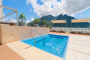 A rooftop pool with a clear view of mountains in the background, surrounded by lounge chairs and potted plants.