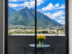 A balcony with a table and chairs, a vase with yellow flowers, and a view of a mountain and town.