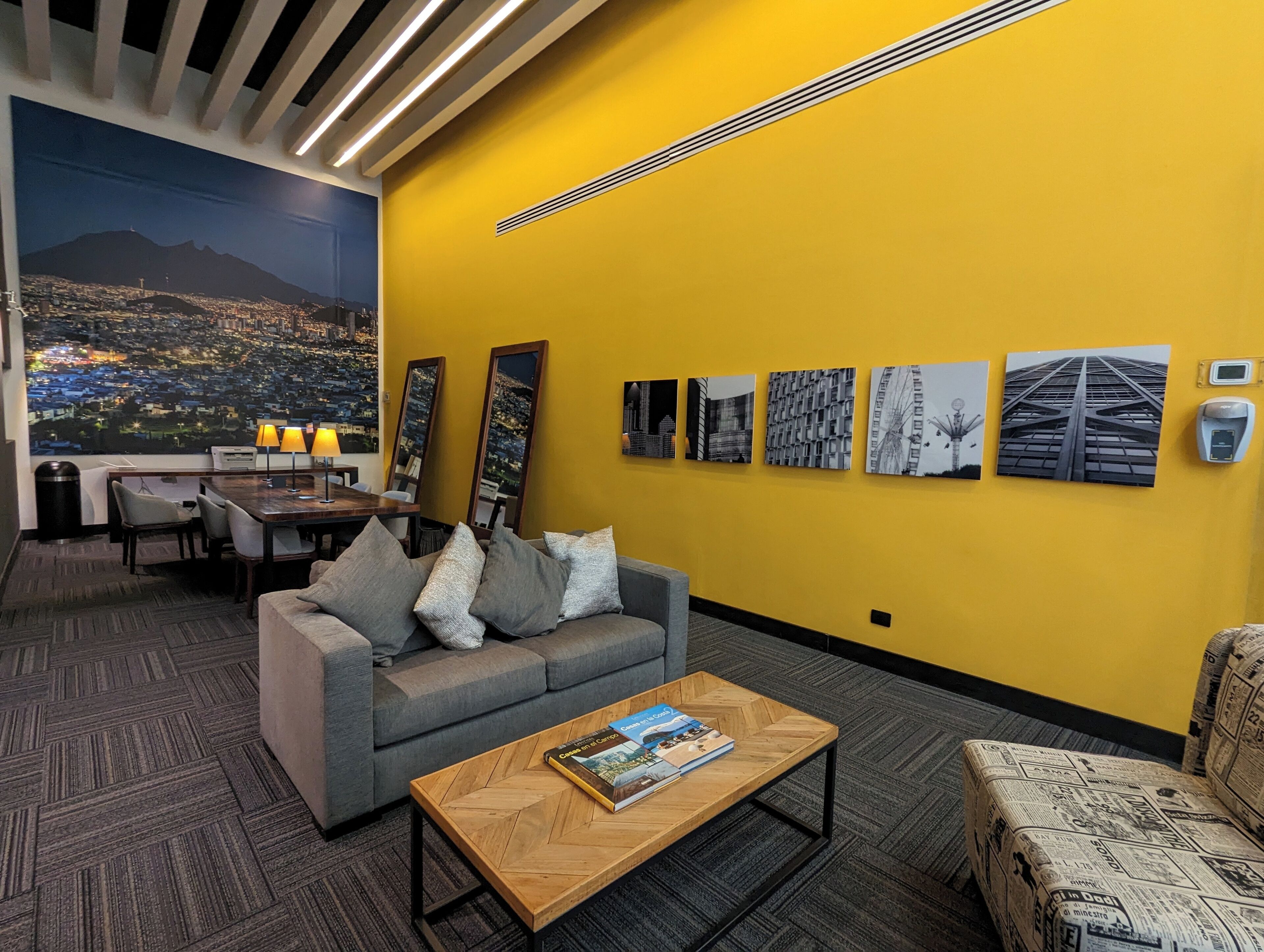 En moderne hotellobby med en gul accentvæg, et stort bybillede som muralmaleri, et spiseområde med bord og stole, samt en sofa med et magasin.
