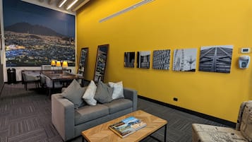 En moderne hotellobby med en gul accentvæg, et stort bybillede som muralmaleri, et spiseområde med bord og stole, samt en sofa med et magasin.