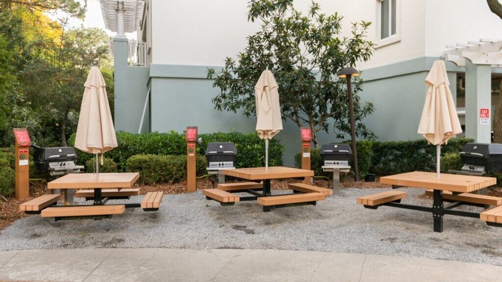 An outdoor seating area with picnic tables and barbecue grills, surrounded by greenery and a building.
