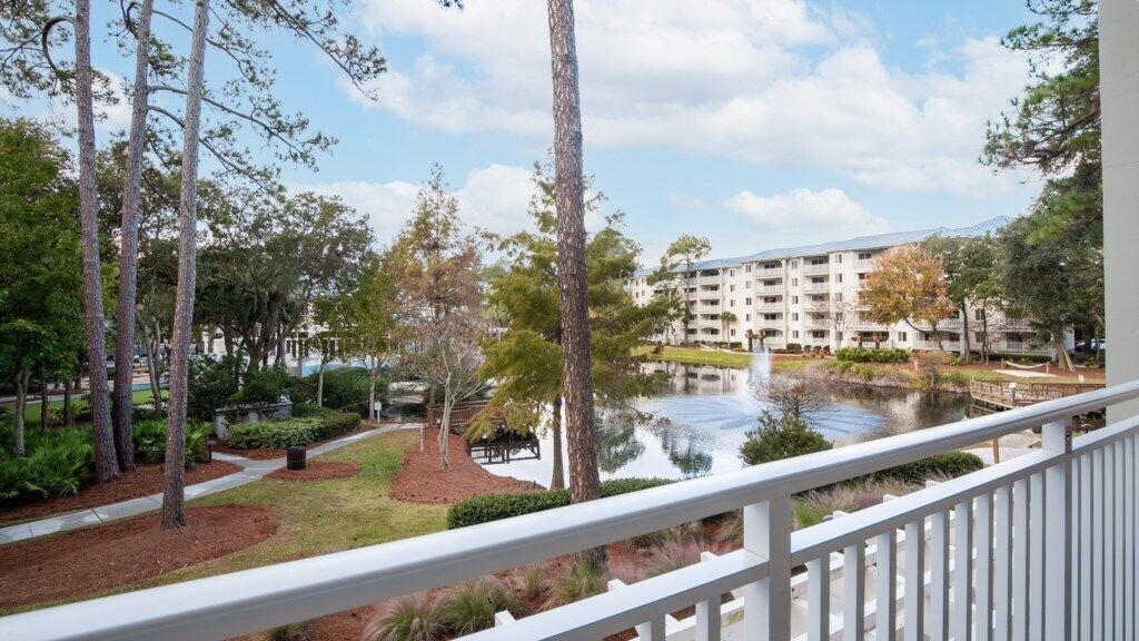 A balcony with a view of a pond, trees, and a multi-story building in the background.