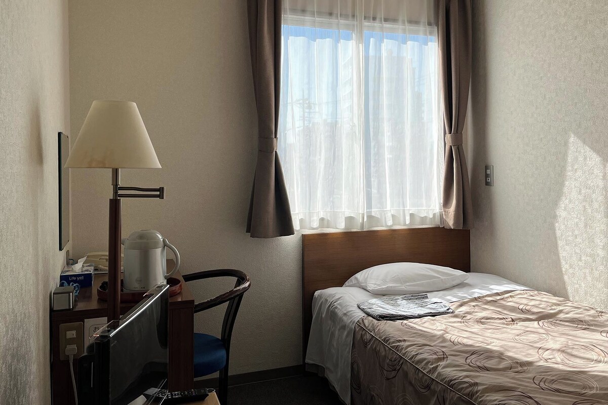 Une chambre d’hôtel avec un lit, un bureau avec une lampe, une chaise et une fenêtre avec des rideaux.