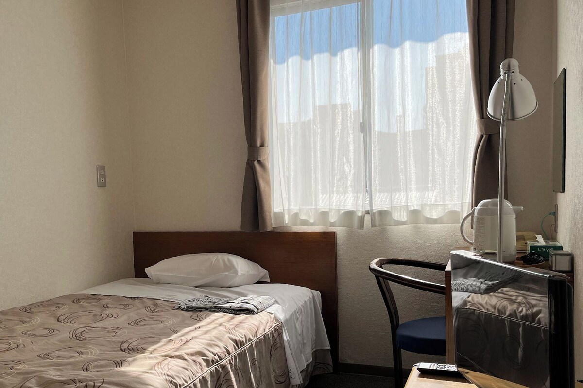 Une petite chambre d’hôtel avec un lit simple, un bureau avec une chaise, une table de chevet et une fenêtre avec des rideaux légers.