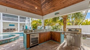 Una cocina al aire libre con una parrilla empotrada, una piscina y un área de estar cubierta.