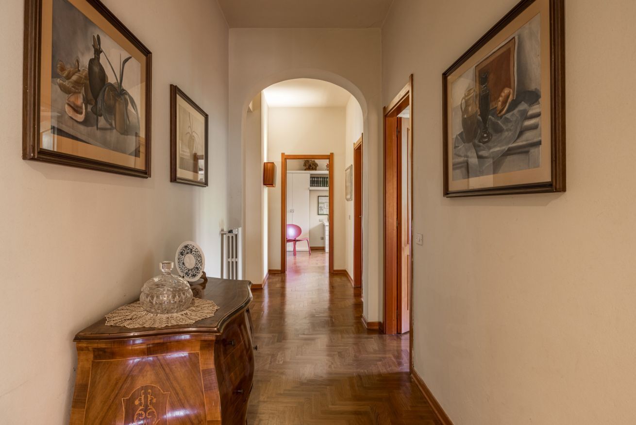 Un pasillo con piso de madera, obras de arte enmarcadas en las paredes, un aparador de madera con superficie de vidrio y un arco de entrada a otra habitación.