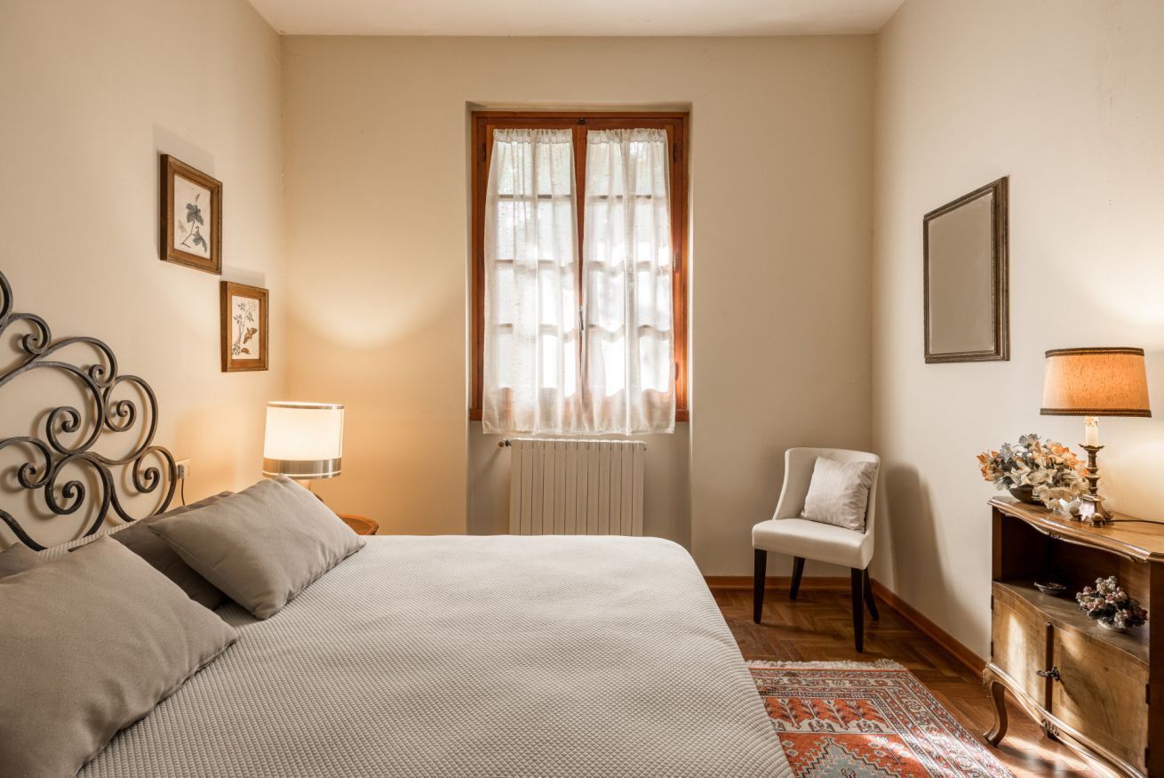 A bedroom with a bed, a chair, a nightstand, a lamp, and a window with curtains.